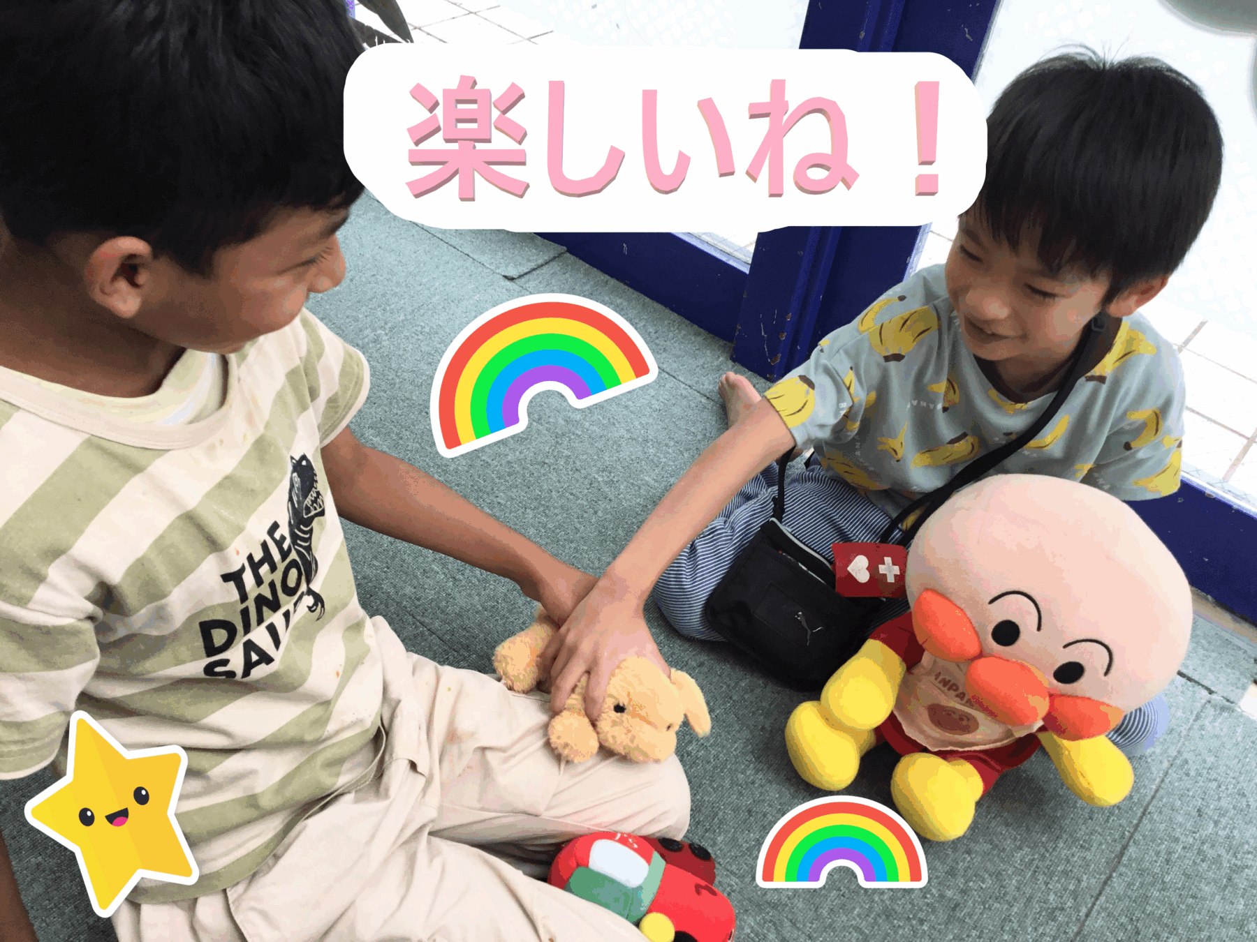 Two kids sitting on a carpet, smiling and holding a small stuffed toy while colorful rainbow stickers and a large pink text banner saying '楽しいね！' appear above them.