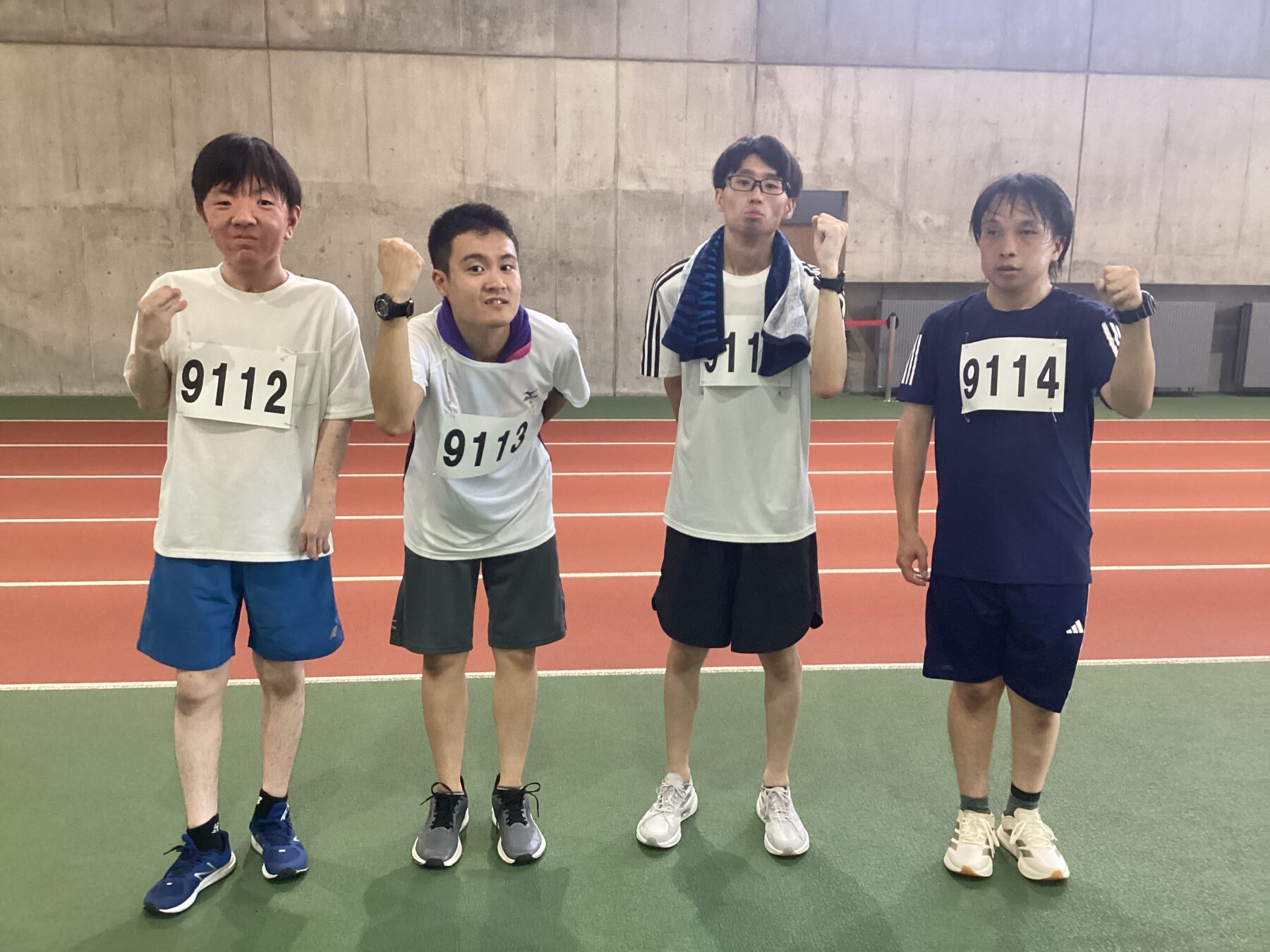 Four men in athletic gear posing on an indoor track, fists raised, with race bibs 9112–9114 visible on chests.