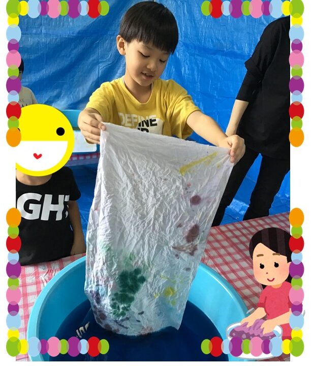 Young boy in a yellow shirt holds up a wrinkled white cloth smeared with green, purple, and yellow paint over a blue tub at a kids' art station.