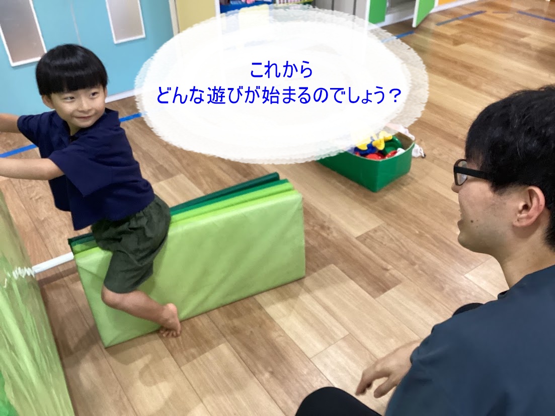 Child climbs over a green foam block in a colorful playroom while an adult watches nearby; speech bubble reads, 'これからどんな遊びが始まるのでしょう?'