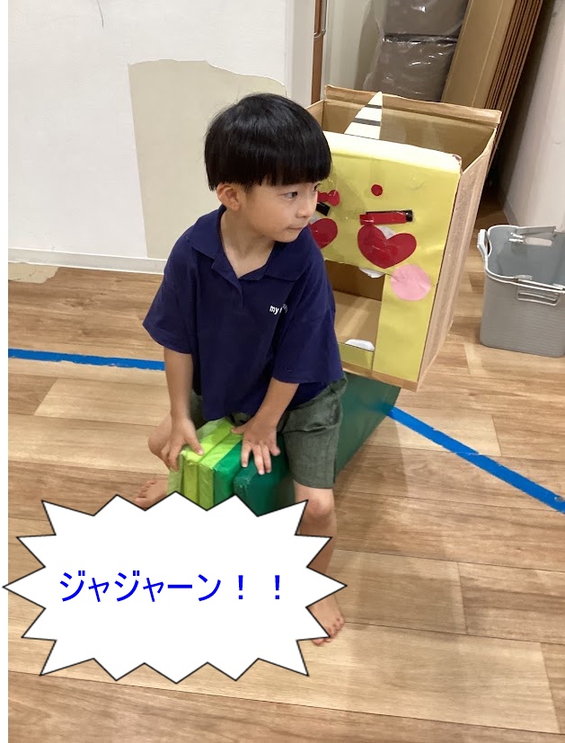 Young child in a blue polo sits on green blocks wearing a homemade cardboard robot costume (yellow head, cone antenna); speech bubble reads ジャジャーン!!