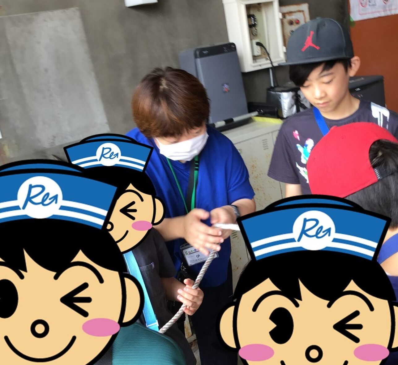 Group of kids wearing blue lanyards and cartoon hats, handling a rope in a workshop with an adult supervisor nearby.