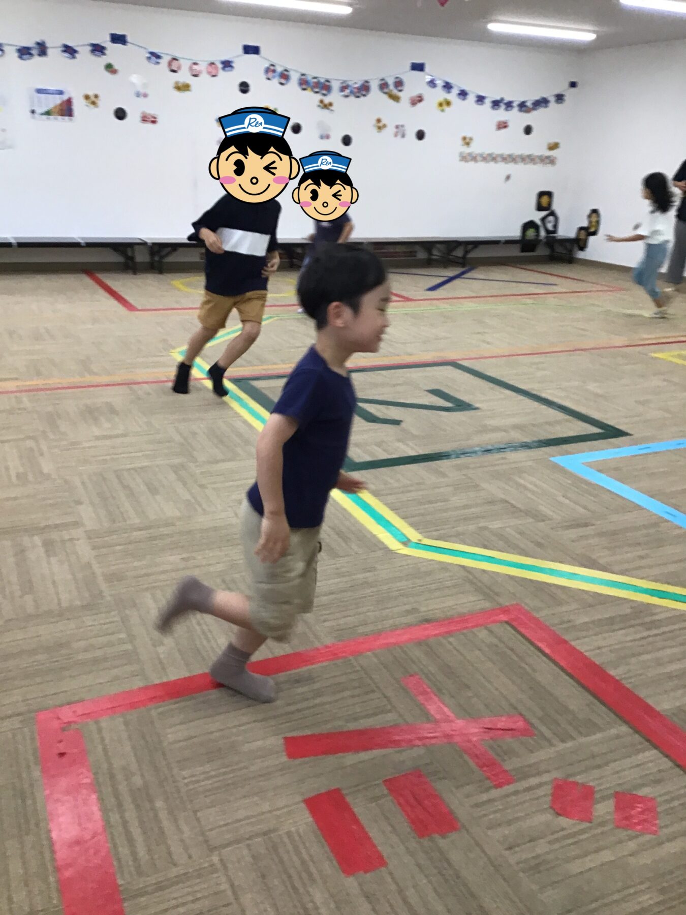 Children running in a gym with colorful taped lines on the floor and wall decorations in the background; two kids in focus wearing dark shirts and shorts, cartoon face stickers obscuring them, others nearby.