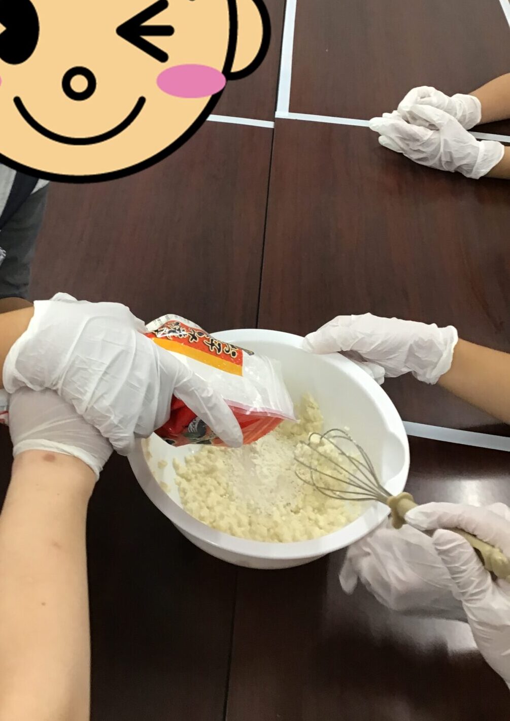 Child wearing gloves stirs flour in a white bowl with a whisk on a table during a baking activity in a classroom setting.