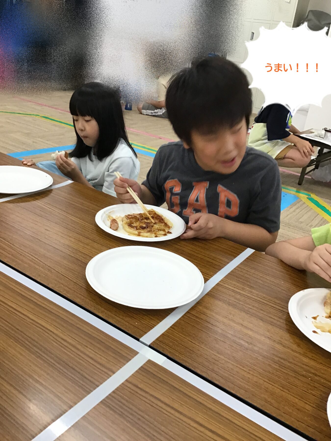 Two children sit at a long wooden table eating pizza; girl on the left looks down at her slice while the boy on the right leans forward with chopsticks. Plates are in front, background slightly blurred.