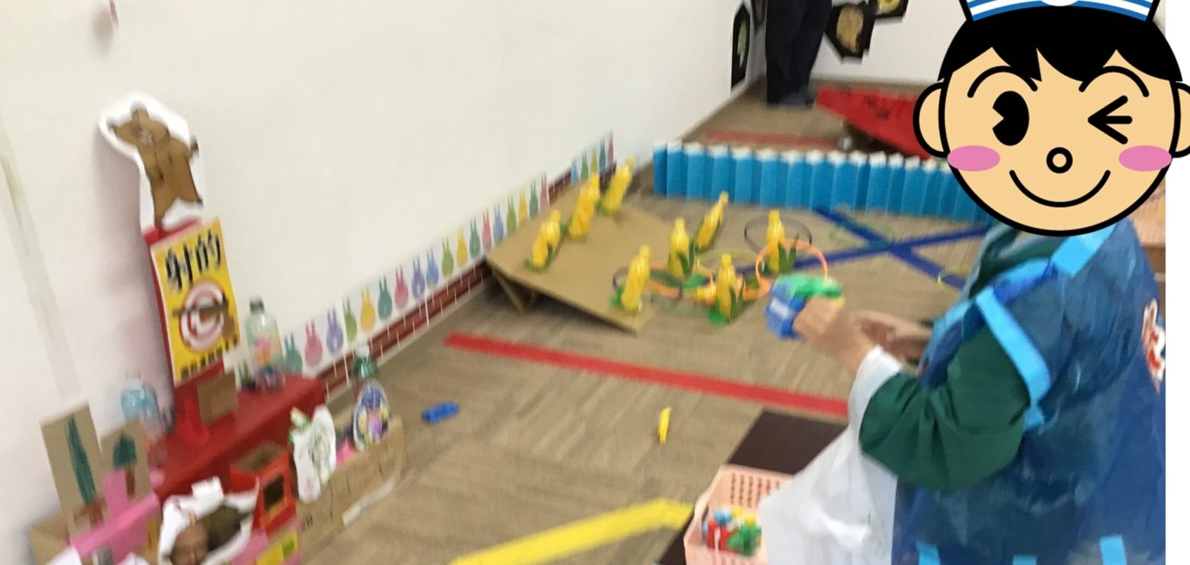 Child in a blue cape and colorful outfit stands at a pretend play setup in a classroom with yellow toy figures on a ramp.