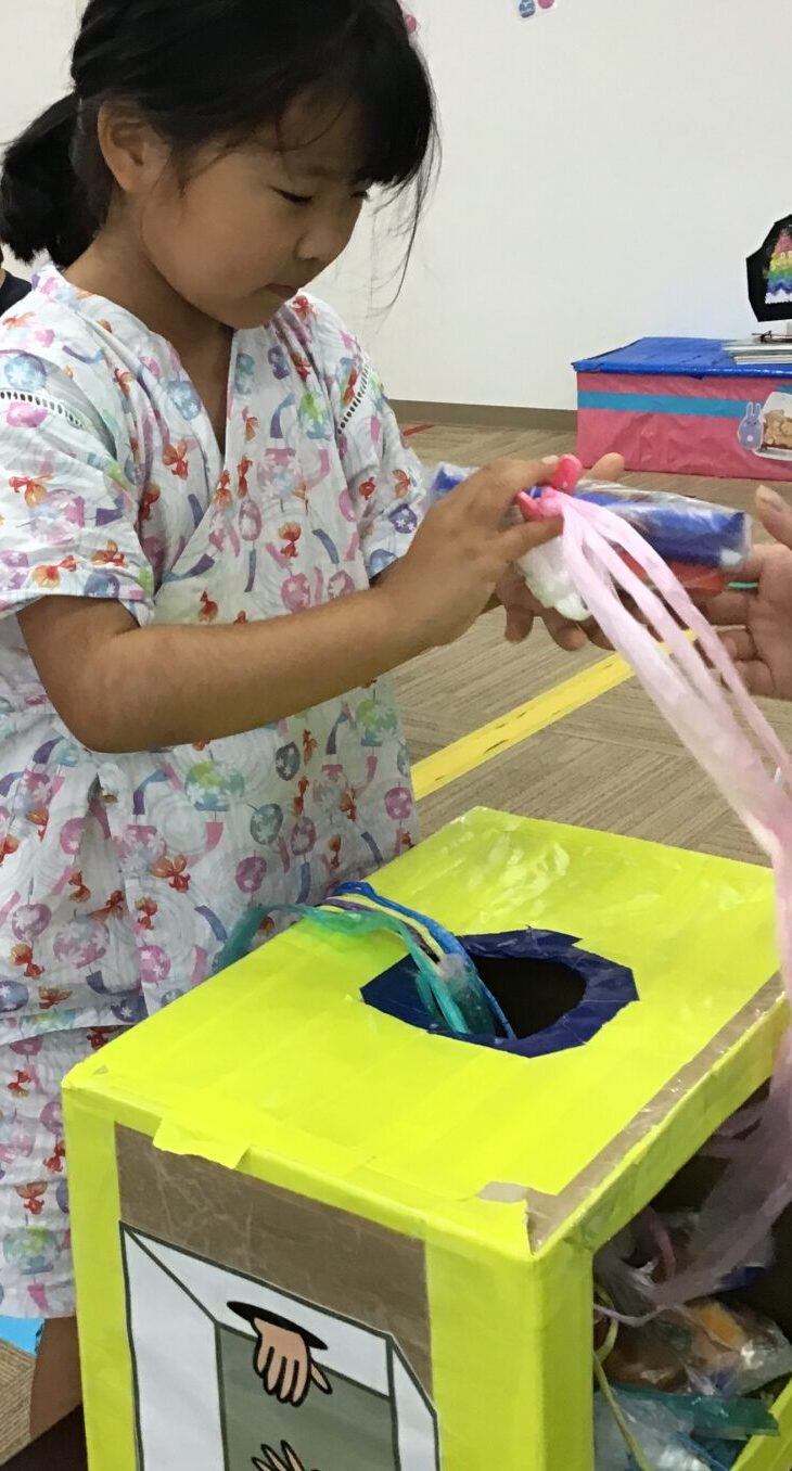 A young girl in a pastel-patterned outfit places pink ribbons into a bright yellow art box during a classroom craft activity, with other children in the background.
