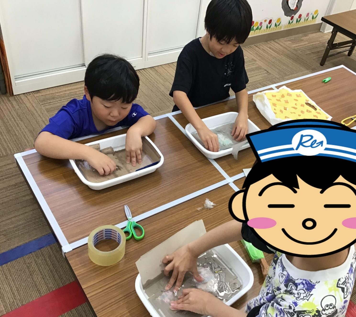 Two children sit at a table, hands in shallow trays of muddy water during a classroom sensory activity outside the frame.