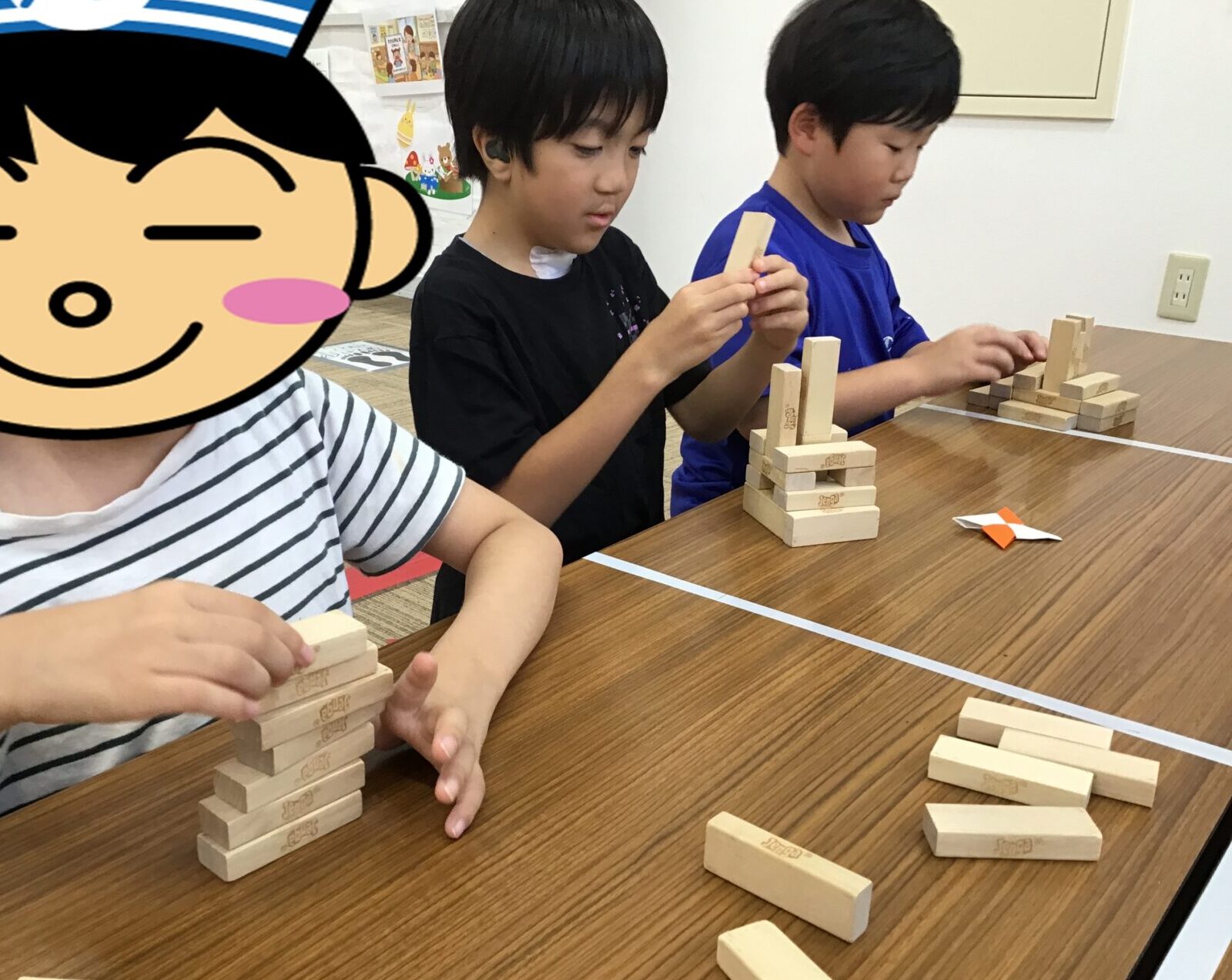 Three children play Jenga at a wooden table, stacking and removing blocks in a classroom setting.