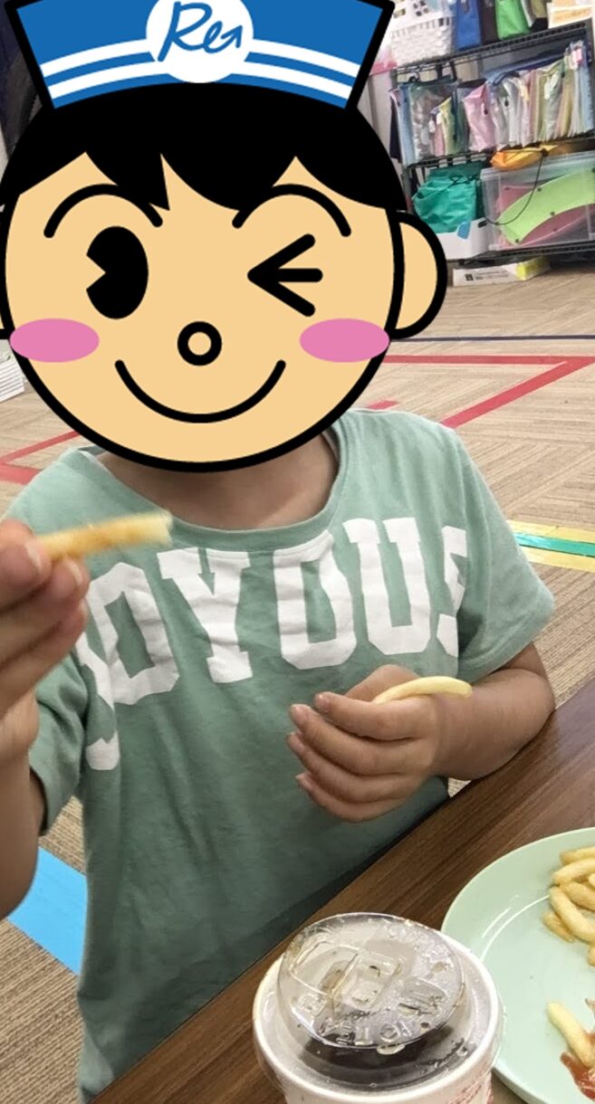 Child in a mint-green shirt sits at a table in a store or cafe, holding a French fry toward the camera; a drink cup and plate of fries are visible.