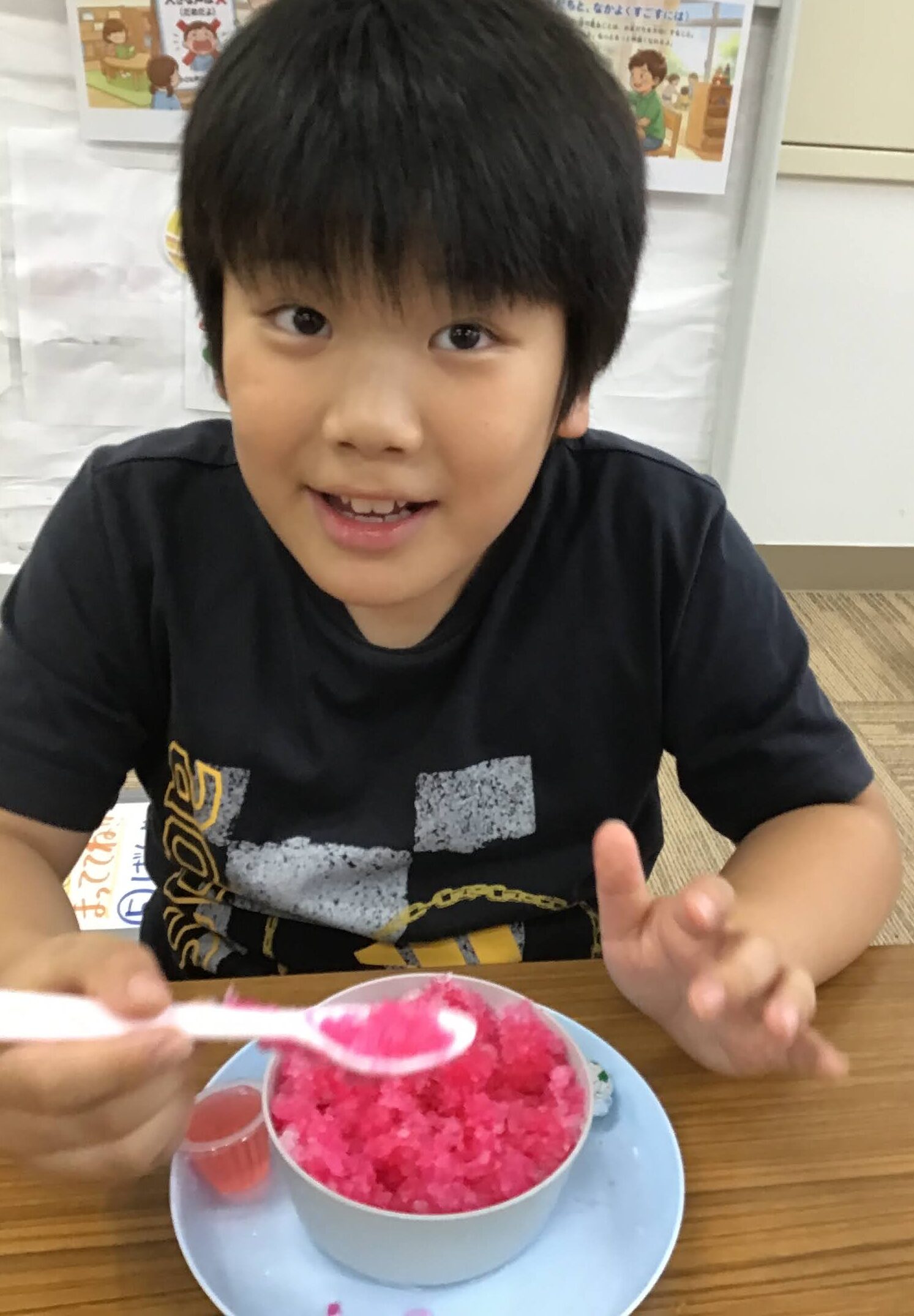 Young boy at a table with a pink shaved-ice dessert in a blue bowl, holding a spoon in front of him in a classroom setting