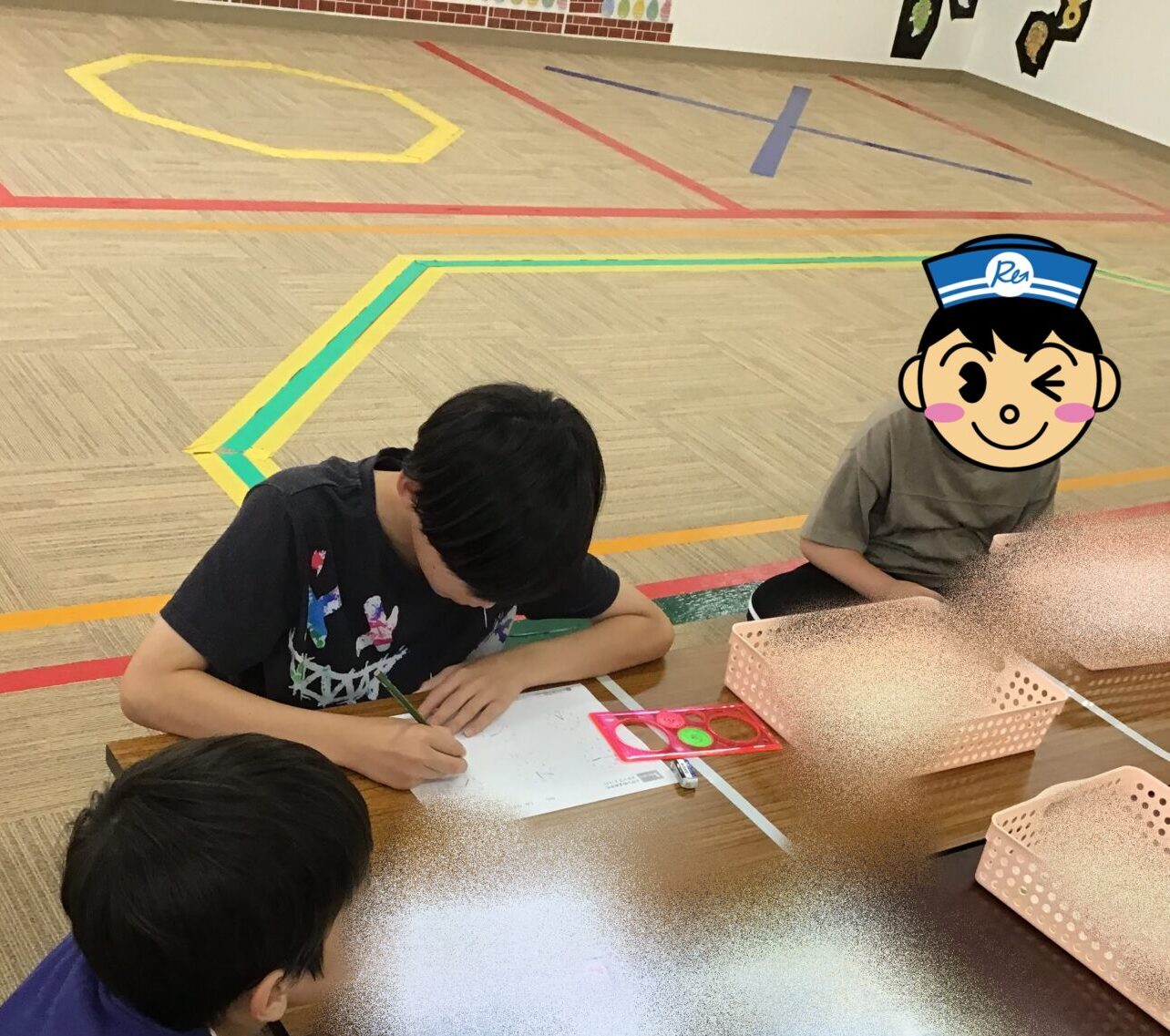 Three children sit at a low table drawing on white paper with pencils, while plastic baskets sit nearby in a classroom gym. Behind them are colorful floor markings on a wooden floor.