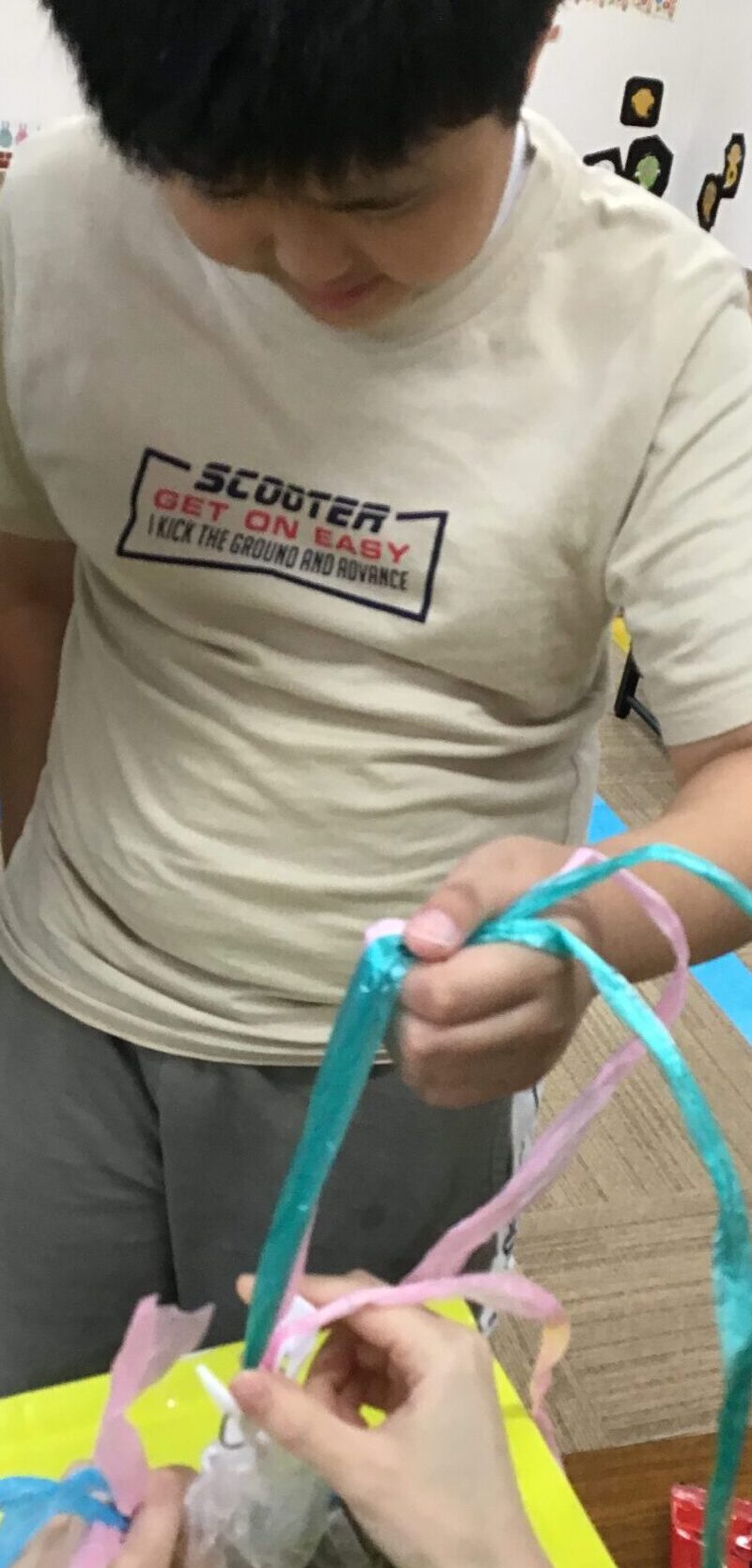Child in a white shirt ties colorful ribbons during a craft activity at a table with others nearby far in the background.