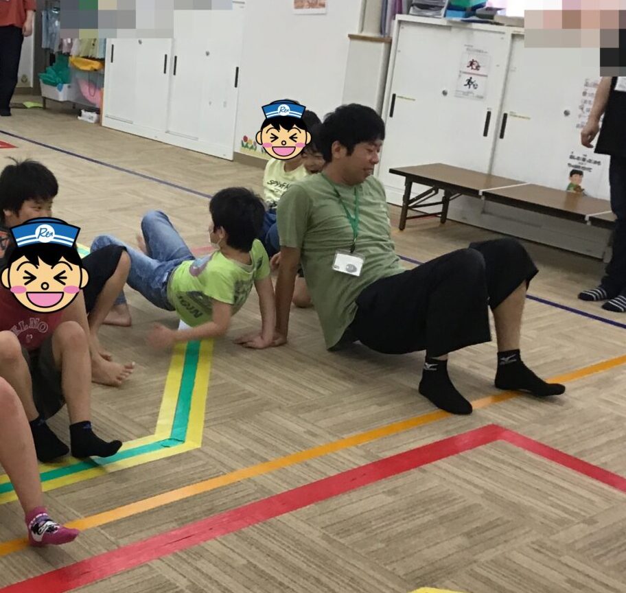 Children seated on a gym floor marked with colorful tape lines, indoors, with adults supervising in the background.