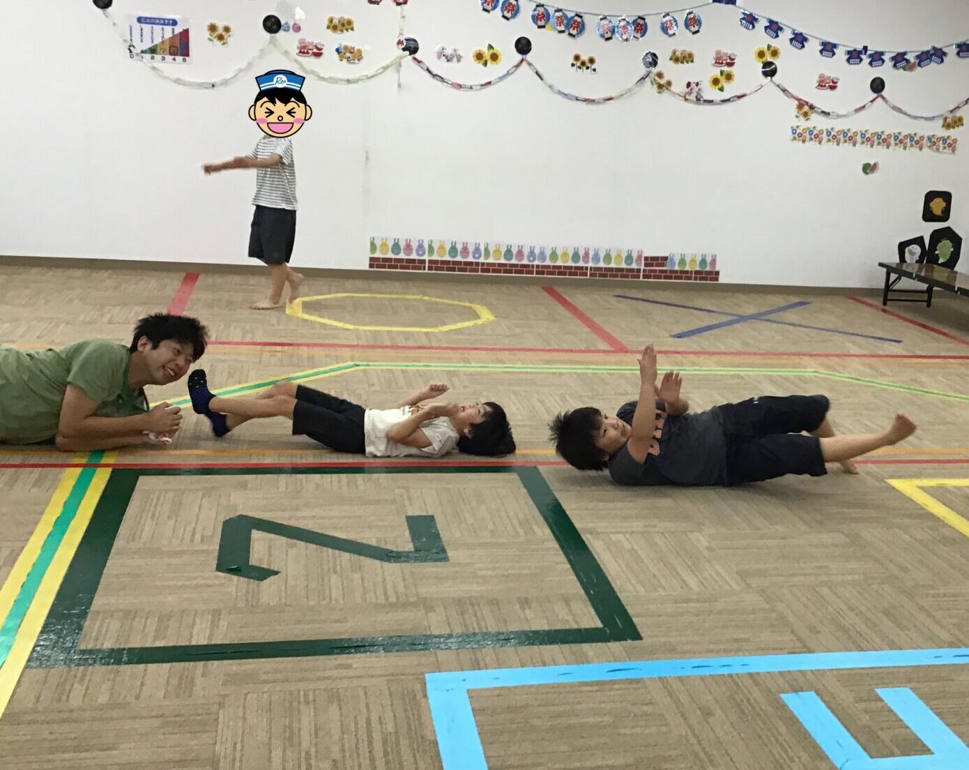 Children lying on their backs doing leg raises on a gym floor while adults supervise; a person stands nearby, and a seated adult with a mask watches on the right, with festive wall decorations in the background.
