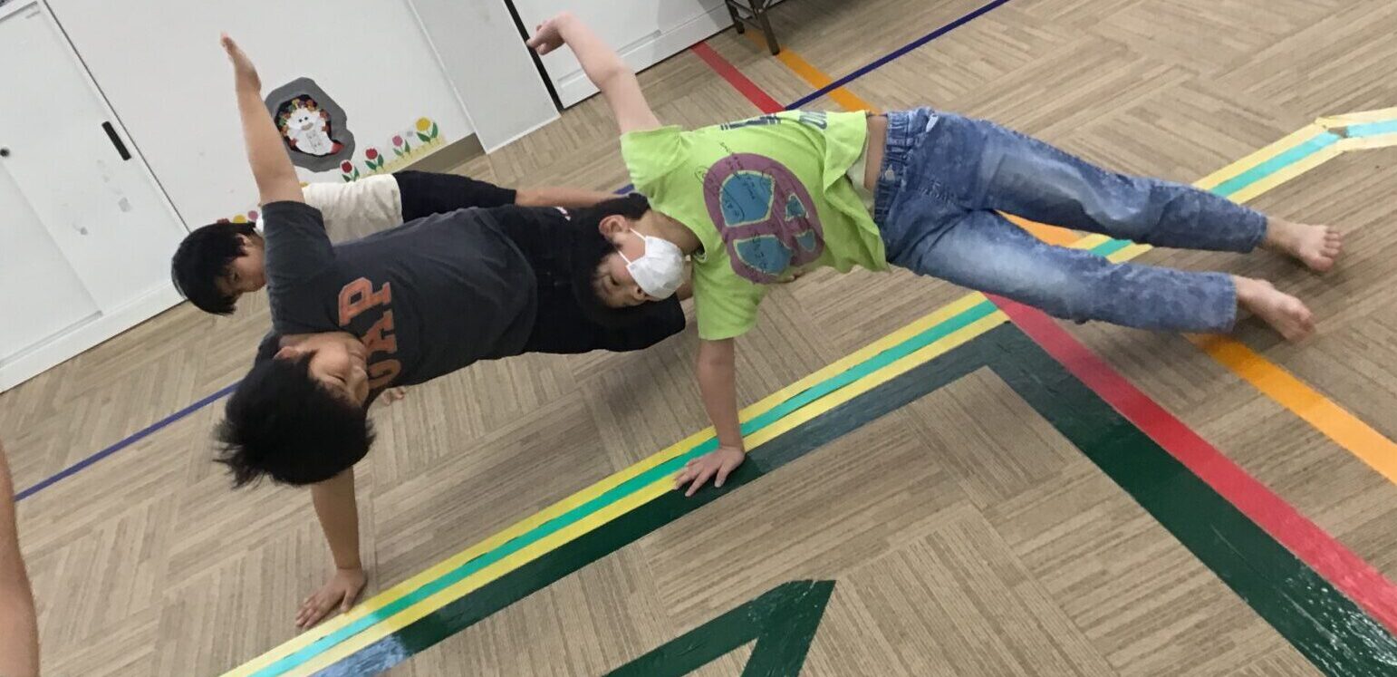 Group of kids in a school gym doing a wheelbarrow walk: one child on hands supported by a partner holding their ankles, others reach with arms for balance.