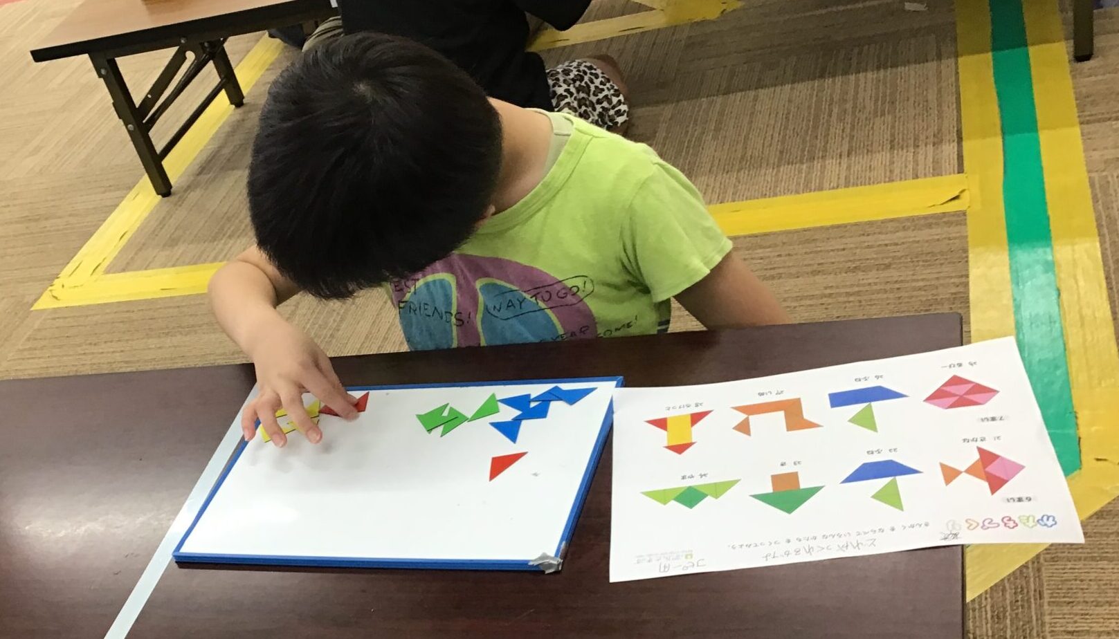 A child in a lime green shirt sits at a table, arranging colorful origami shapes on a workbook in a classroom or gym setting with taped floor lines nearby.