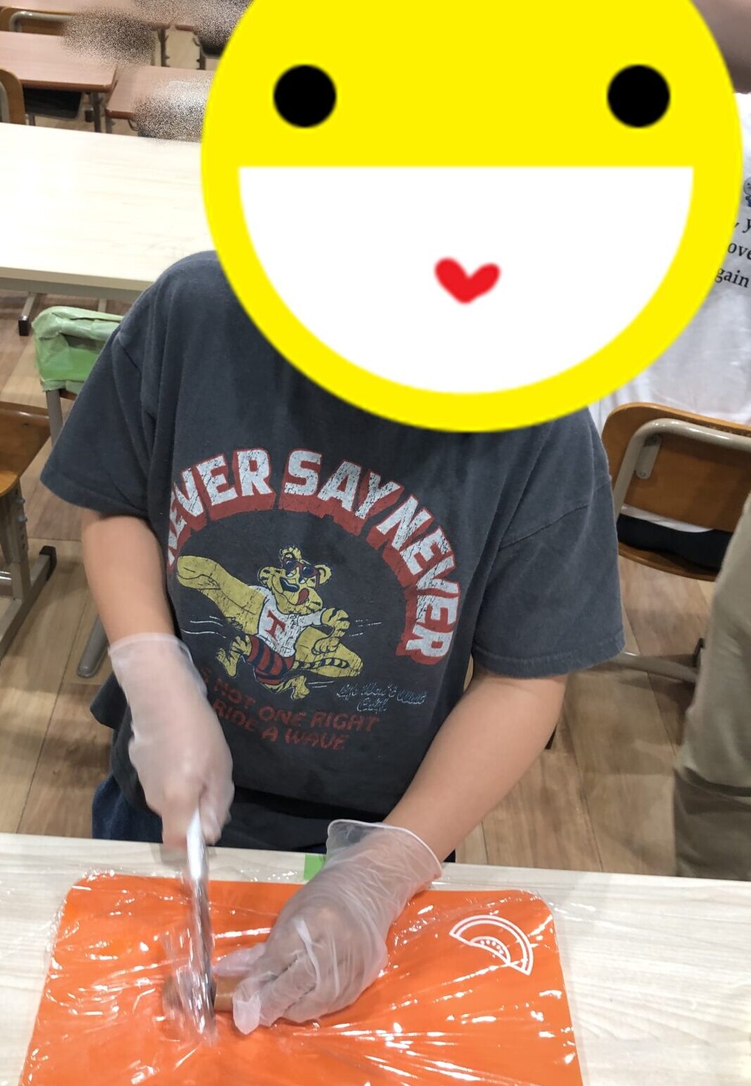 Child wearing gloves and a dark graphic t-shirt cuts bright orange clay or dough on a plastic-covered table in a classroom with desks in the background.