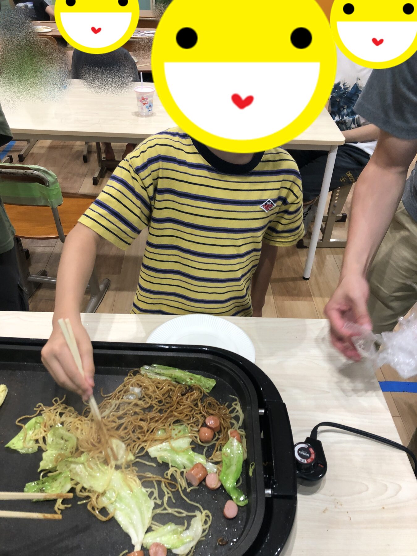 Child in a yellow striped shirt stirs yakisoba with chopsticks on a sizzling griddle, with cabbage and small sausages nearby.