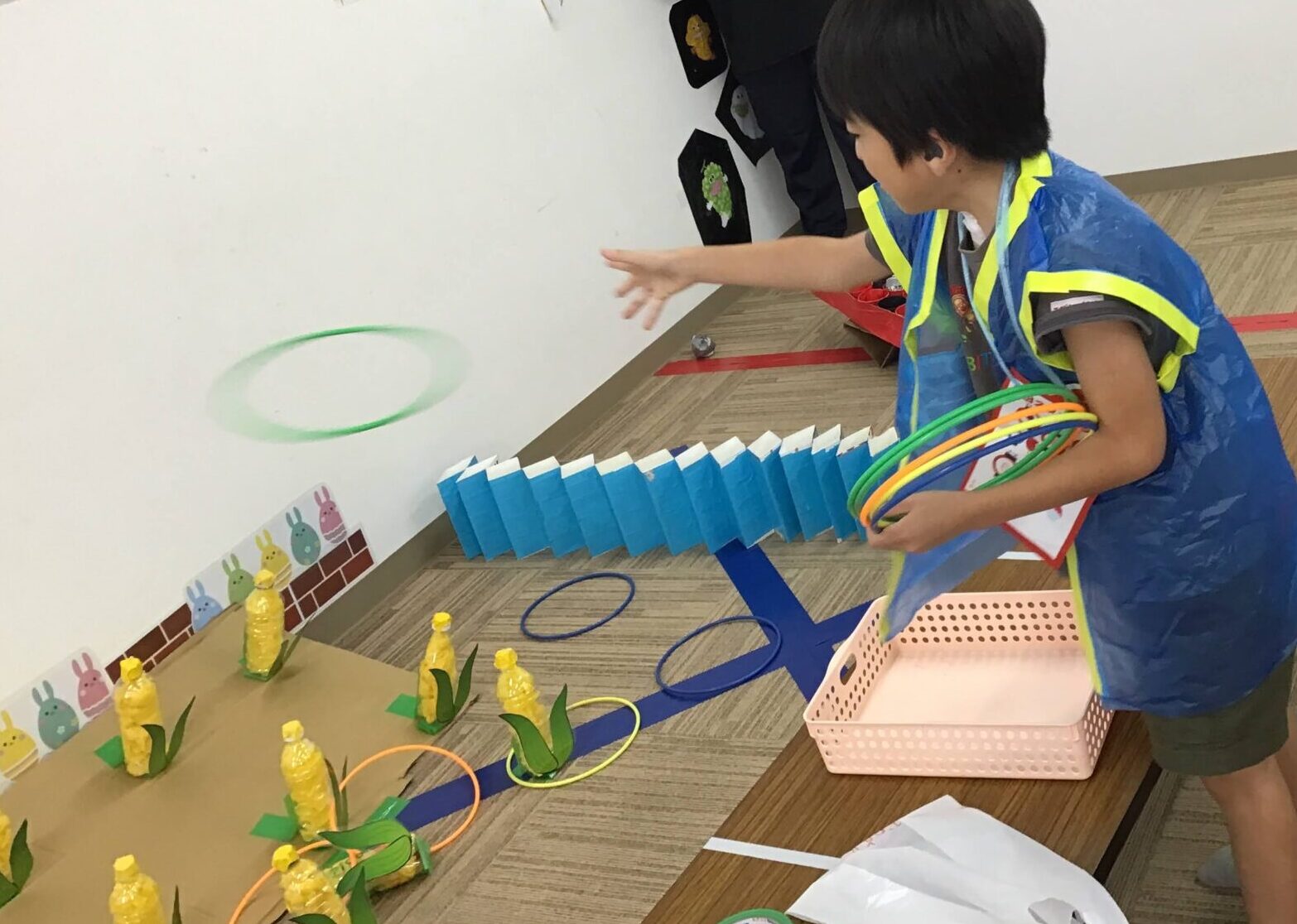 Child in a blue smock tossing hoops toward yellow bottle targets on a game board, with a supervising adult nearby in a classroom setting.
