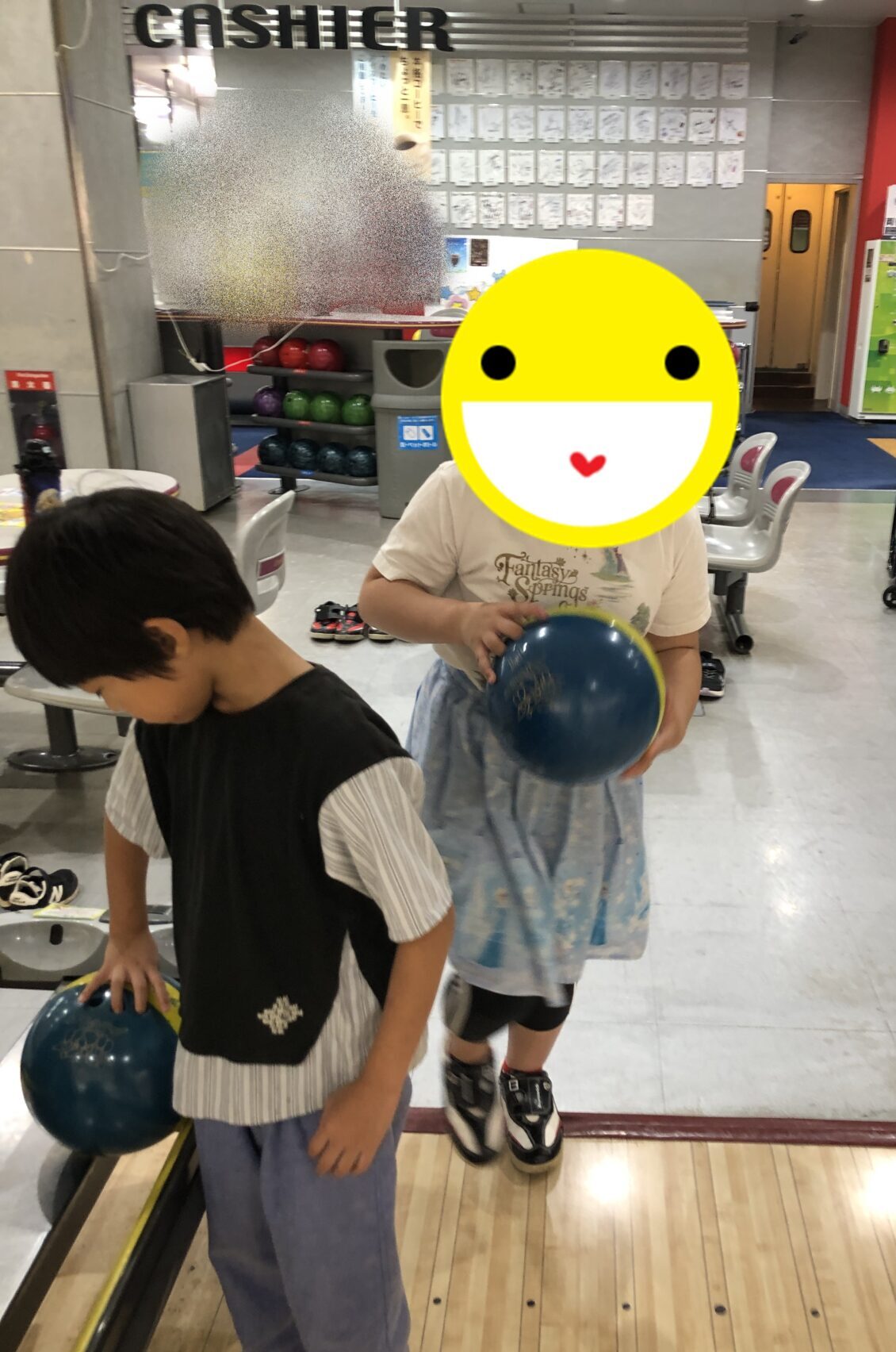 Two kids at a bowling alley preparing to bowl, each holding a blue ball, with a person in a wheelchair and an adult in the background.