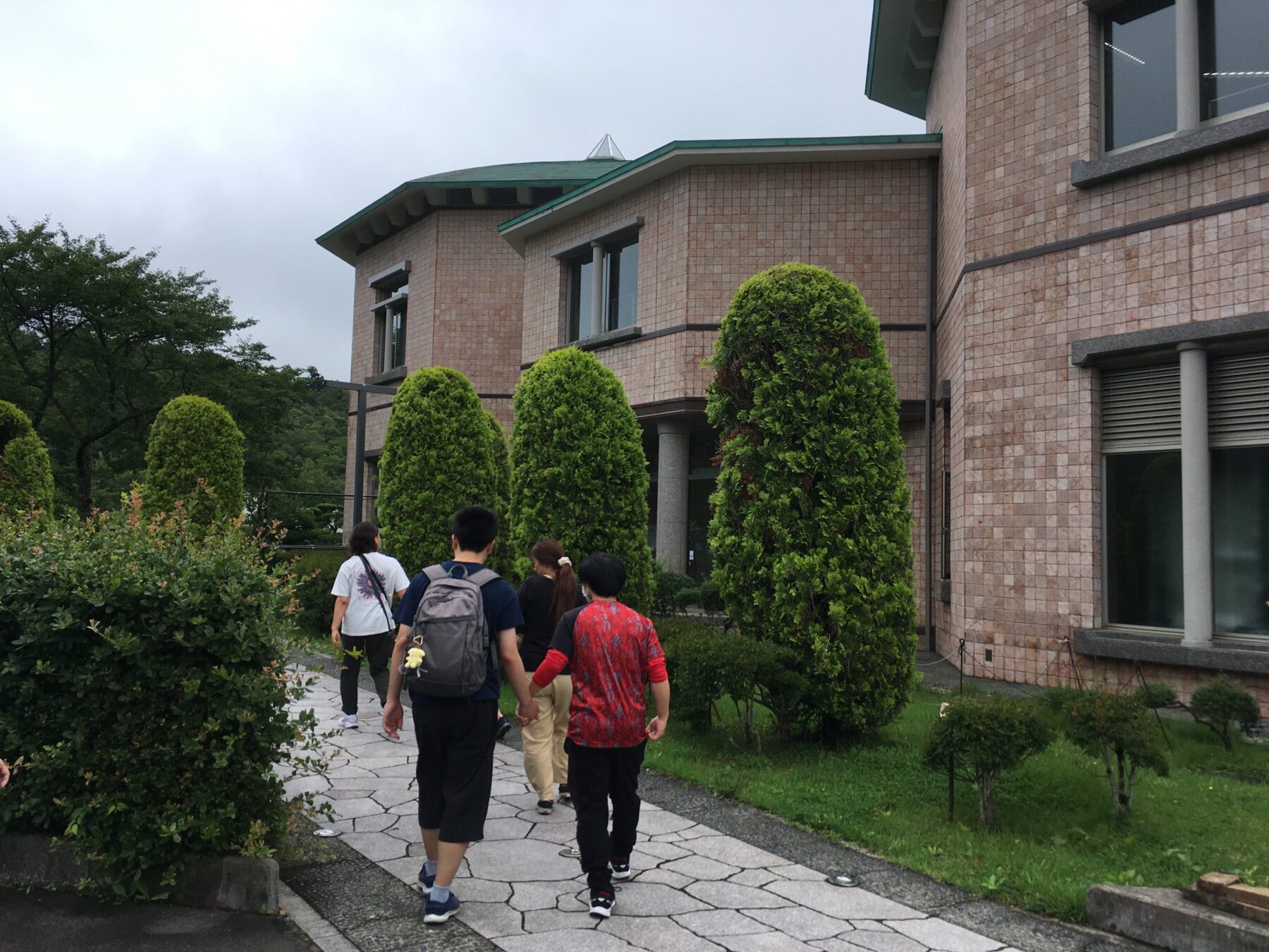 Group of people walking along a stone path beside a tan brick building with a green roof and neatly trimmed shrubs.