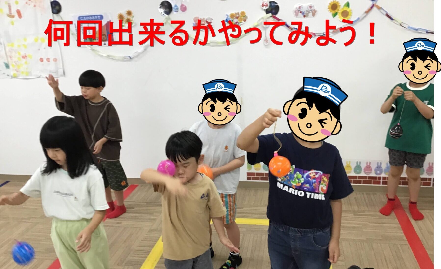 Children in a classroom play with small colorful balls while wearing cartoon face stickers; a red banner with Japanese text hangs on the wall.
