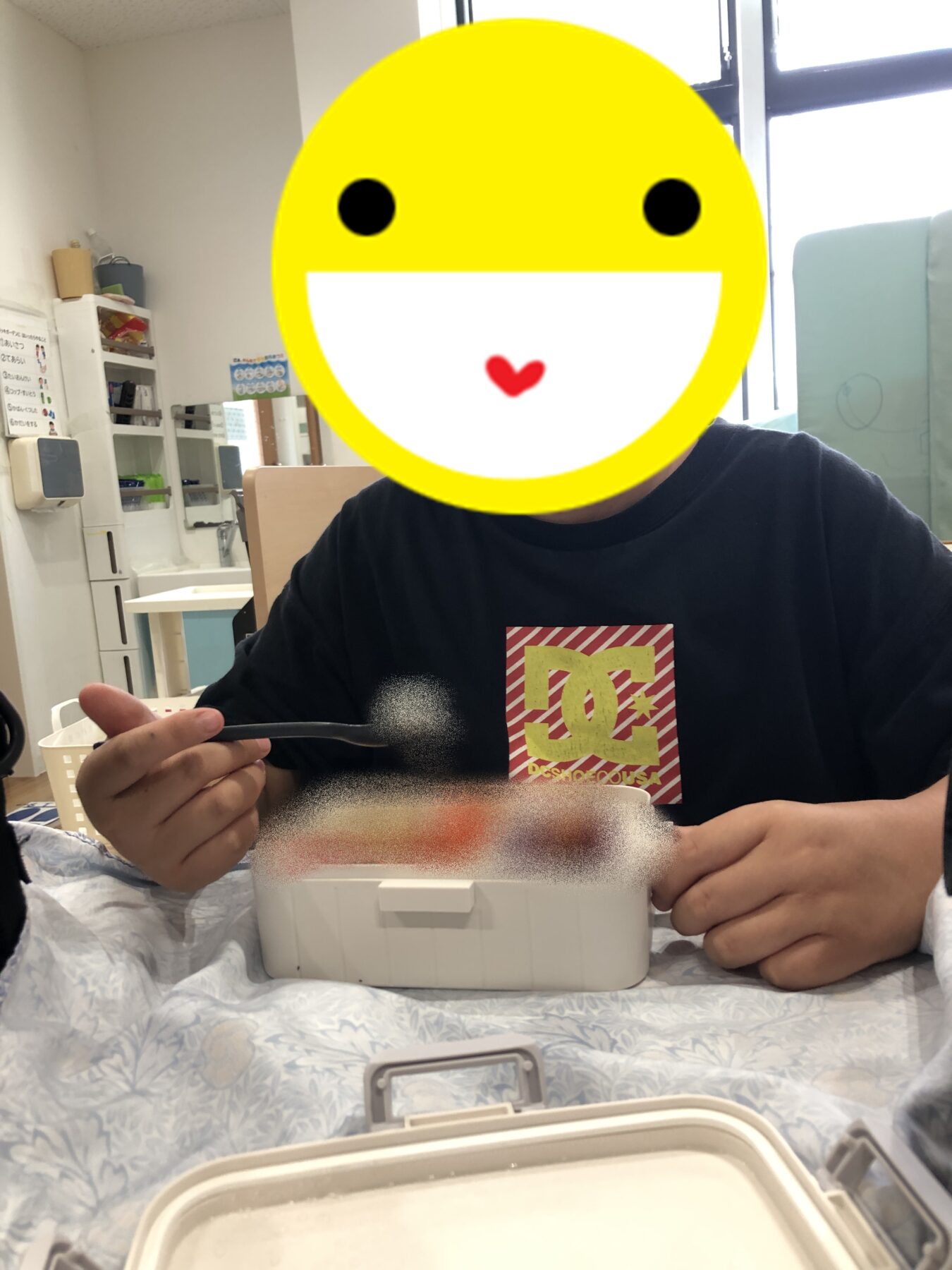 Person seated at a hospital bed tray table, holding a spoon over a white food container; their face is covered by a large yellow smiley sticker.