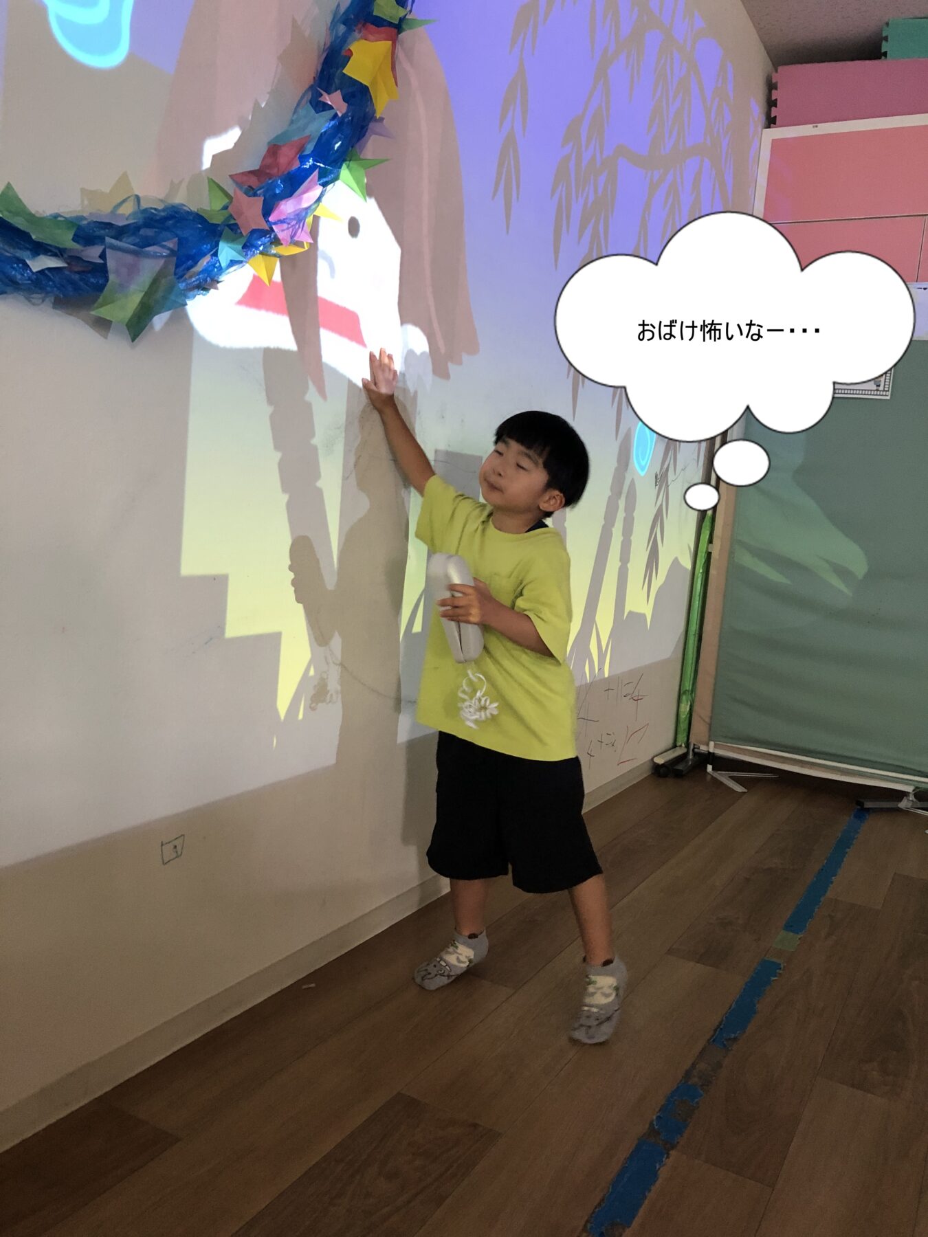 Young child in a lime shirt touches a wall during an interactive projection, with a colorful paper garland overhead and a thought bubble reading 'おばけ怖いな――'.