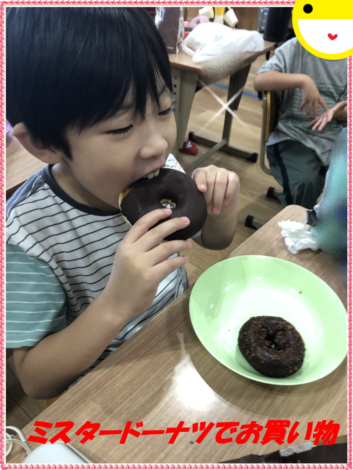 Young child bites into a chocolate-glazed donut at a table, with a green plate and another donut nearby in a classroom or cafe setting.
