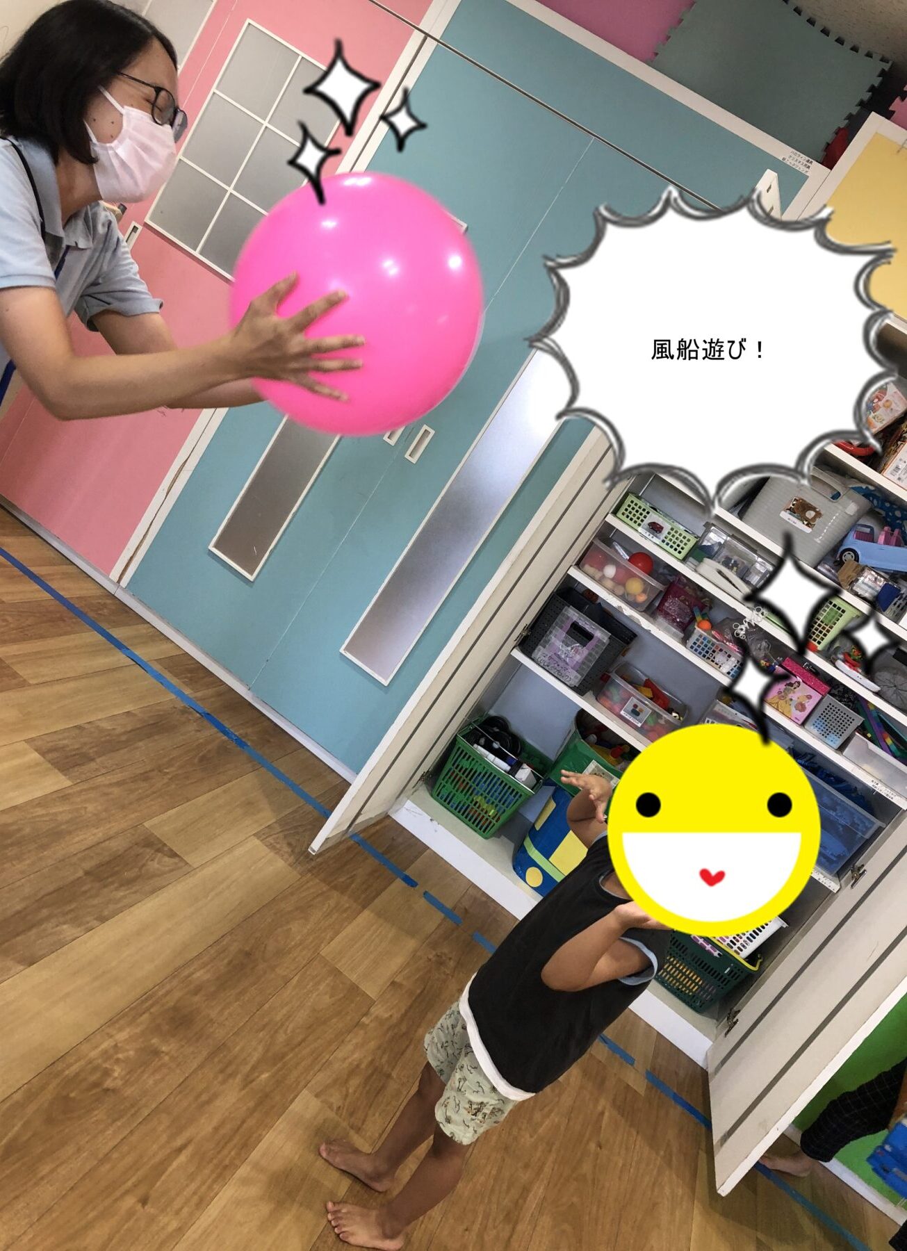 Adult wearing a mask reaches toward a bright pink balloon in a colorful classroom while a playful child watches nearby off-screen (scene with shelves and light blue/pink walls).