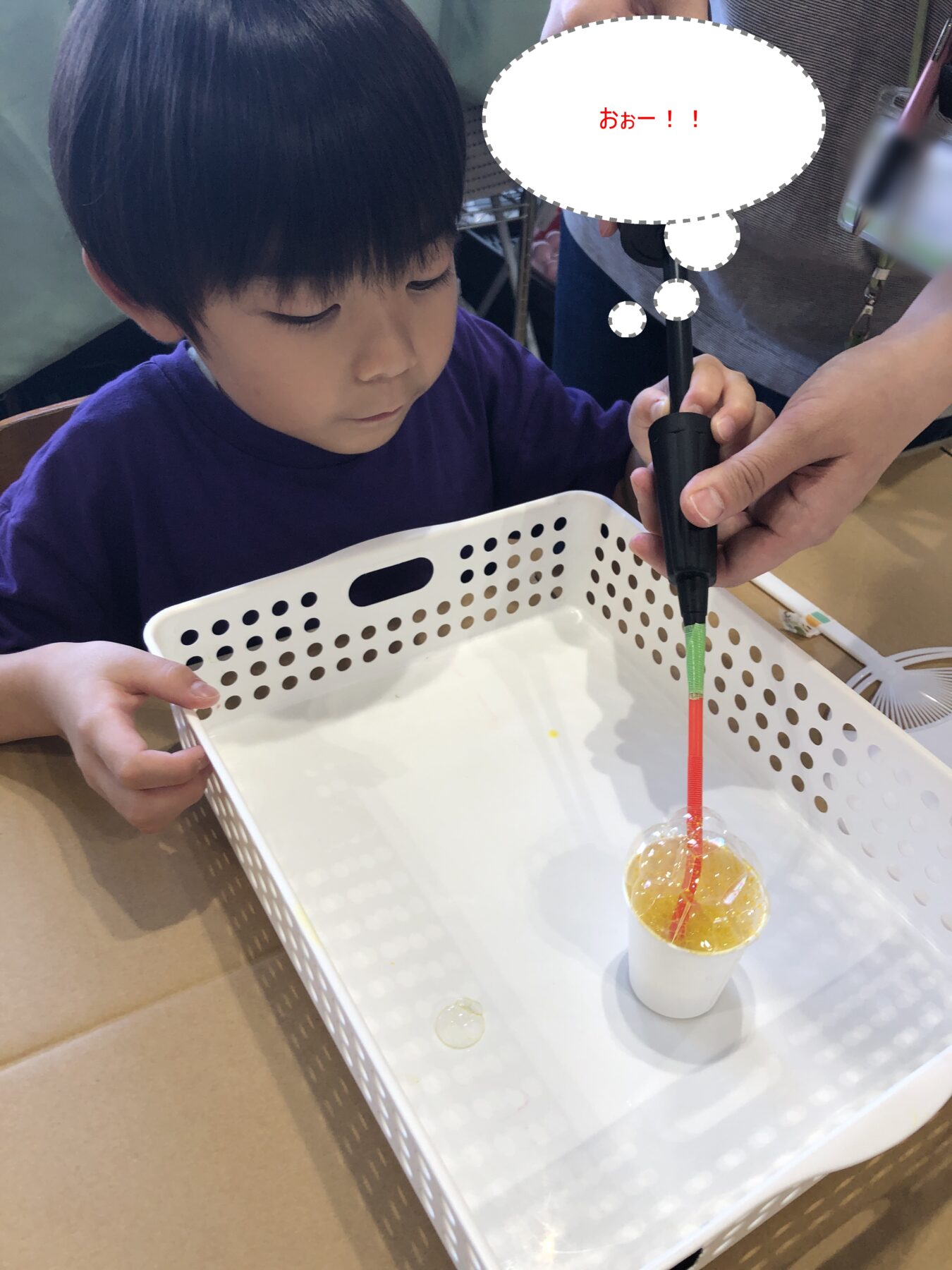 Boy in a purple shirt watches as an adult uses a black handheld tool on a cup with an orange drink and red straw inside a white perforated basket.