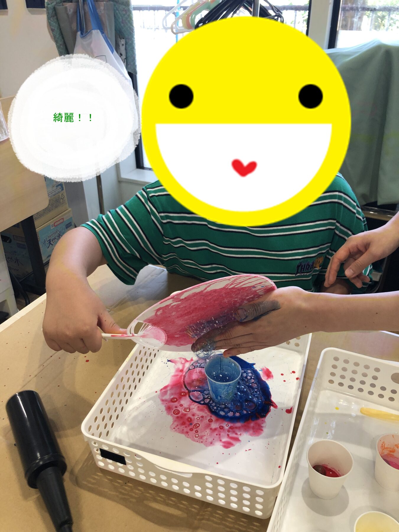 Child wearing a striped shirt paints with pink and blue paint at a table, face covered by a large yellow smiley sticker.