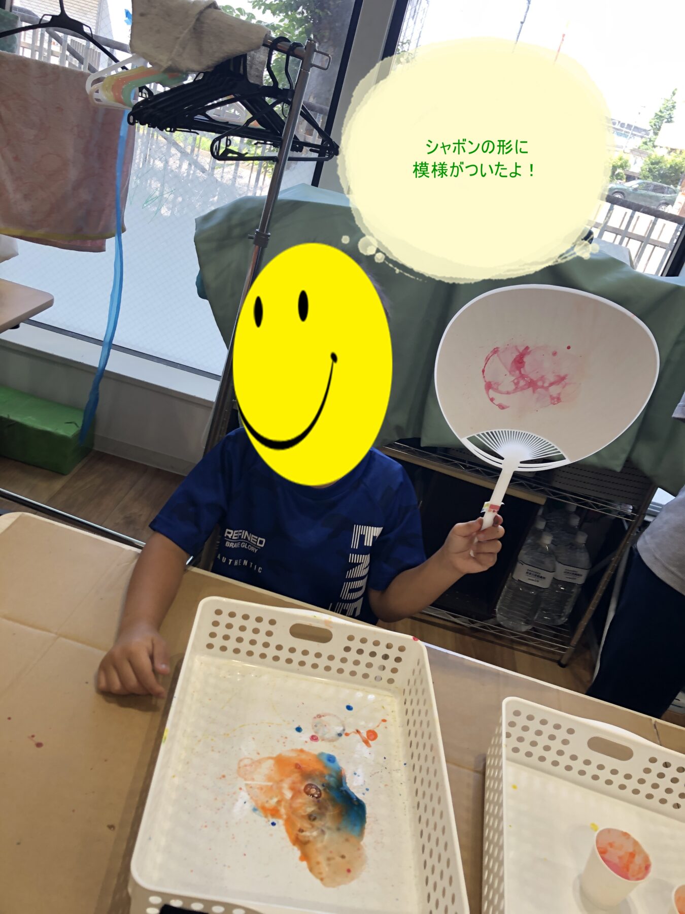 Child seated at a table during a painting activity, with paint in white baskets and a handheld fan showing pink/orange smudges.