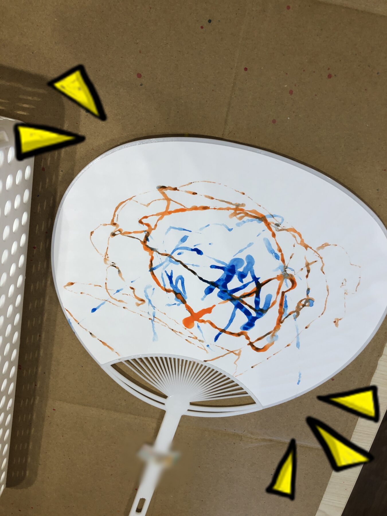 White round hand fan with orange and blue marker scribbles, placed inside a cardboard box (child artwork).
