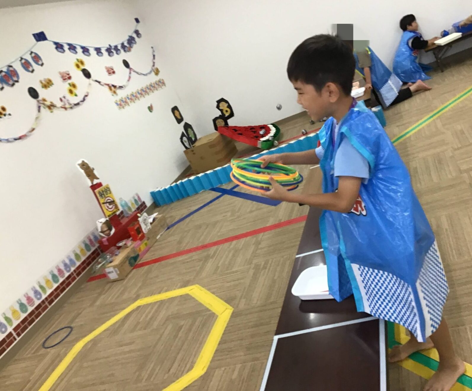 Boy in a blue poncho holding colorful plastic rings at a ring-toss game in a gym with wall decorations behind him.