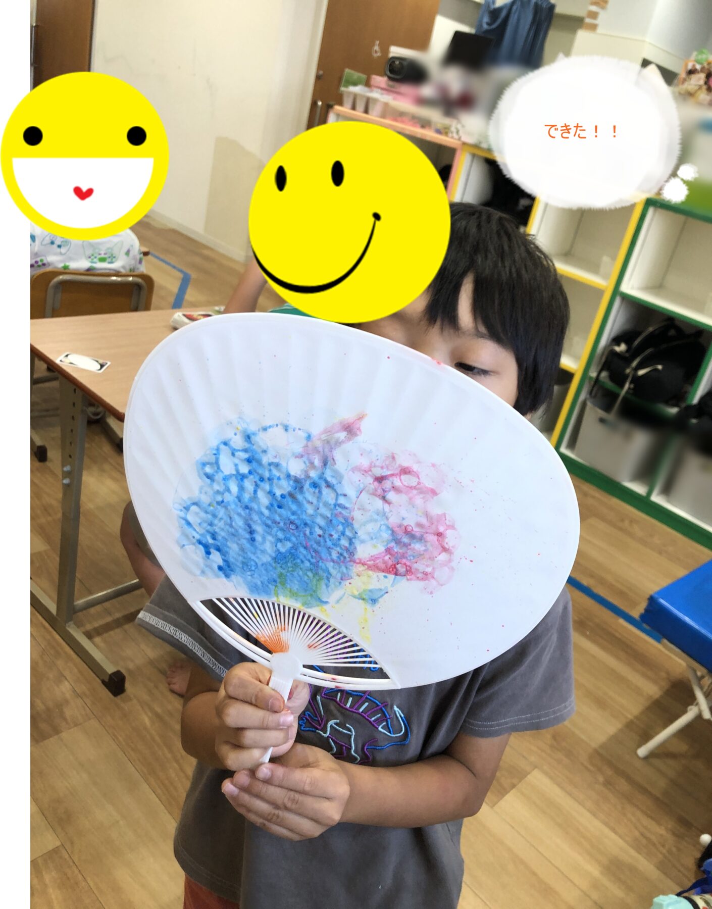 A child in a classroom proudly holding a painted paper fan with blue and pink streaks, smiley faces in the background overlaying the image.