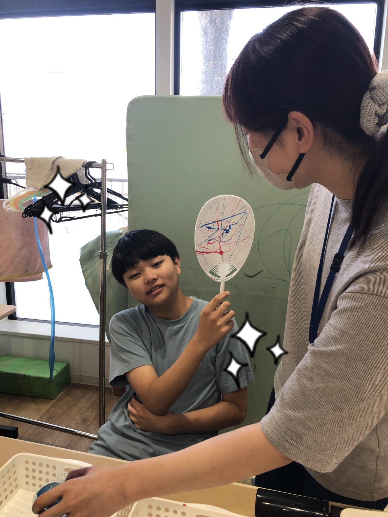 Boy in a blue shirt holds up a white hand fan with colorful scribbles, showing it to a woman wearing a mask in a classroom setting.