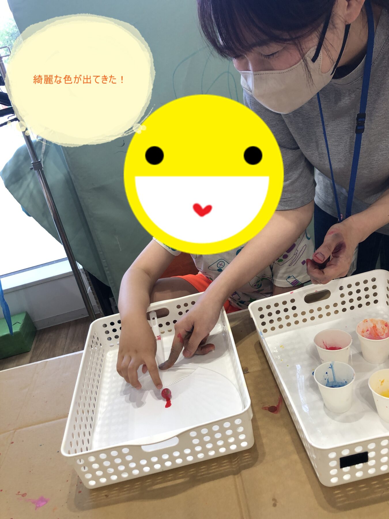 Child and adult wearing a mask painting at a table with white baskets of paint; a large yellow smiley emoji covers the child’s face. Speech bubble reads: 綺麗な色が出てきた!.