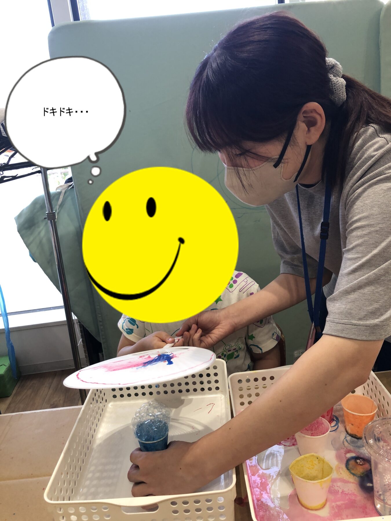 Caregiver helping a child paint at a table with cups of paint and a white plastic caddy nearby in a classroom setting.