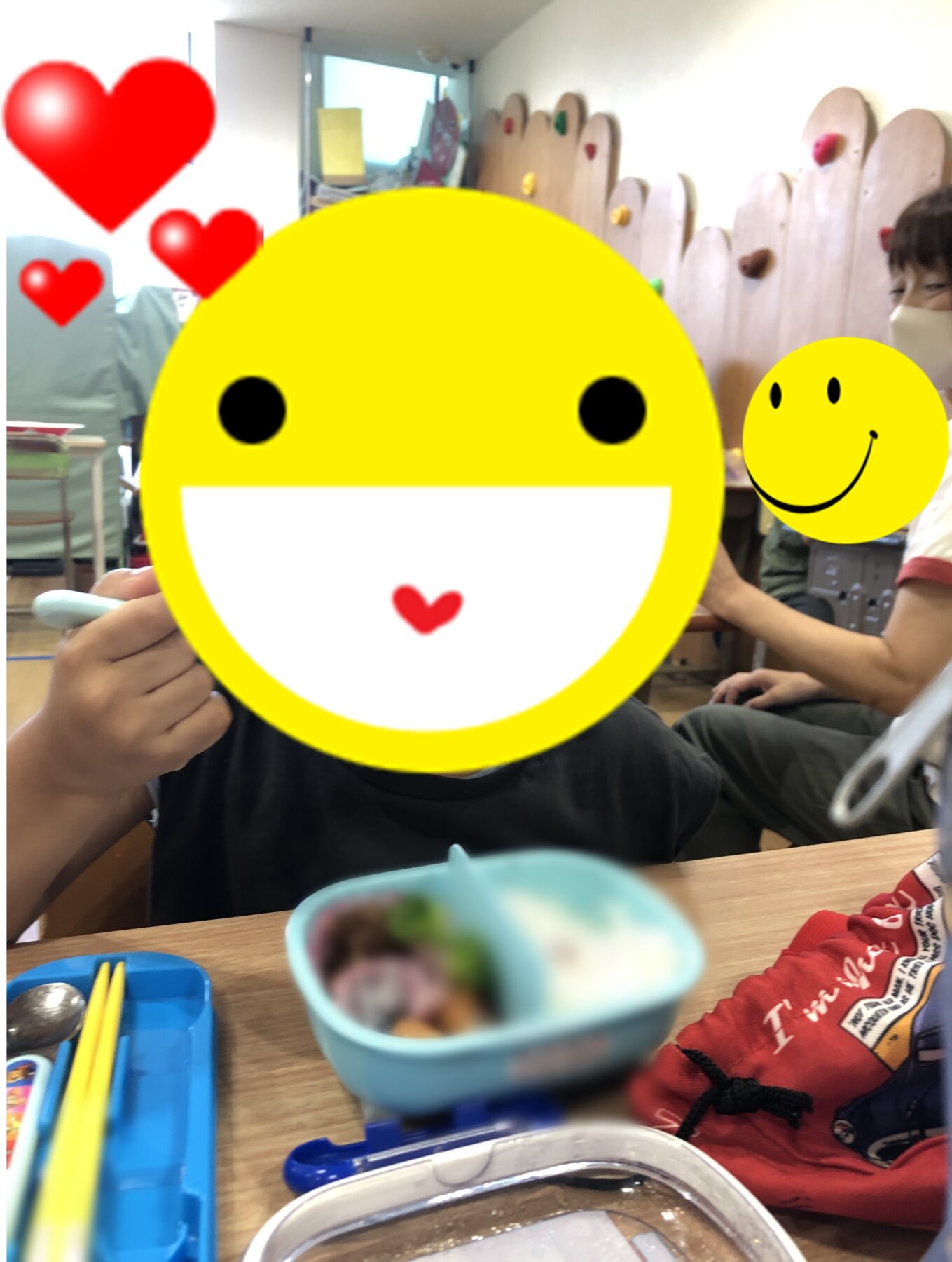 Child at a table eating in a classroom, with a large yellow smiley emoji covering the main person’s face and red heart overlays nearby; another small yellow smiley is visible in the background.