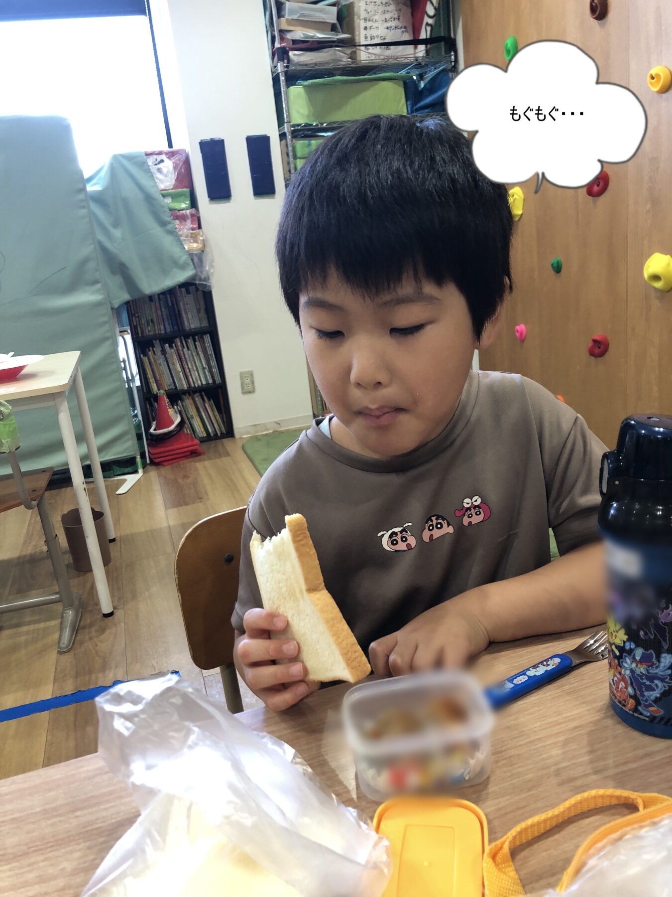 Child sitting at a table, biting into a sandwich in a classroom or activity room setting with shelves in the background.