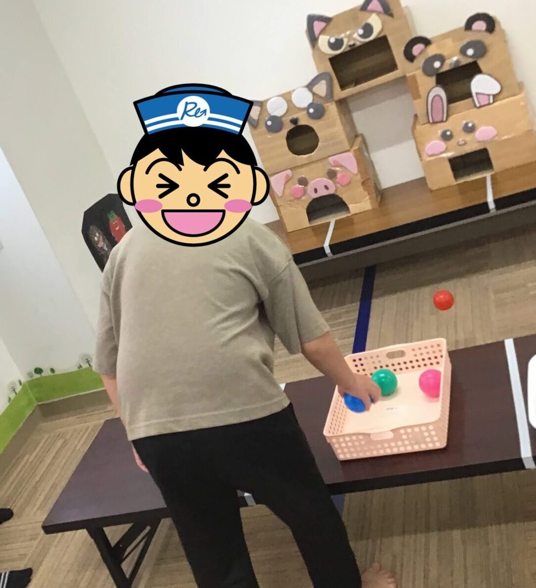 Child wearing a beige shirt plays a ball toss game indoors, reaching for a blue ball from a pink basket, with cardboard animal houses in the background.