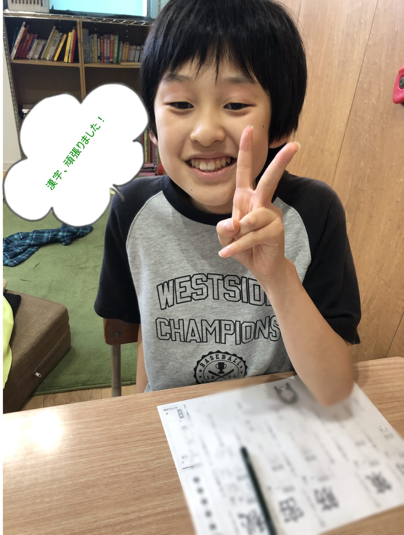 Smiling boy at a desk making a peace sign; a speech bubble on his left contains green Japanese text '漢字、頑張りました!'