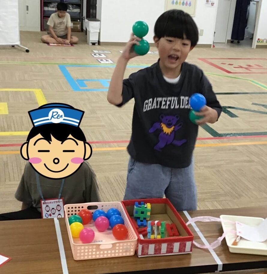Young boy in a classroom holds green balls in one hand and a blue ball in the other, at a table with colorful toys. Behind him, another child sits on the floor.