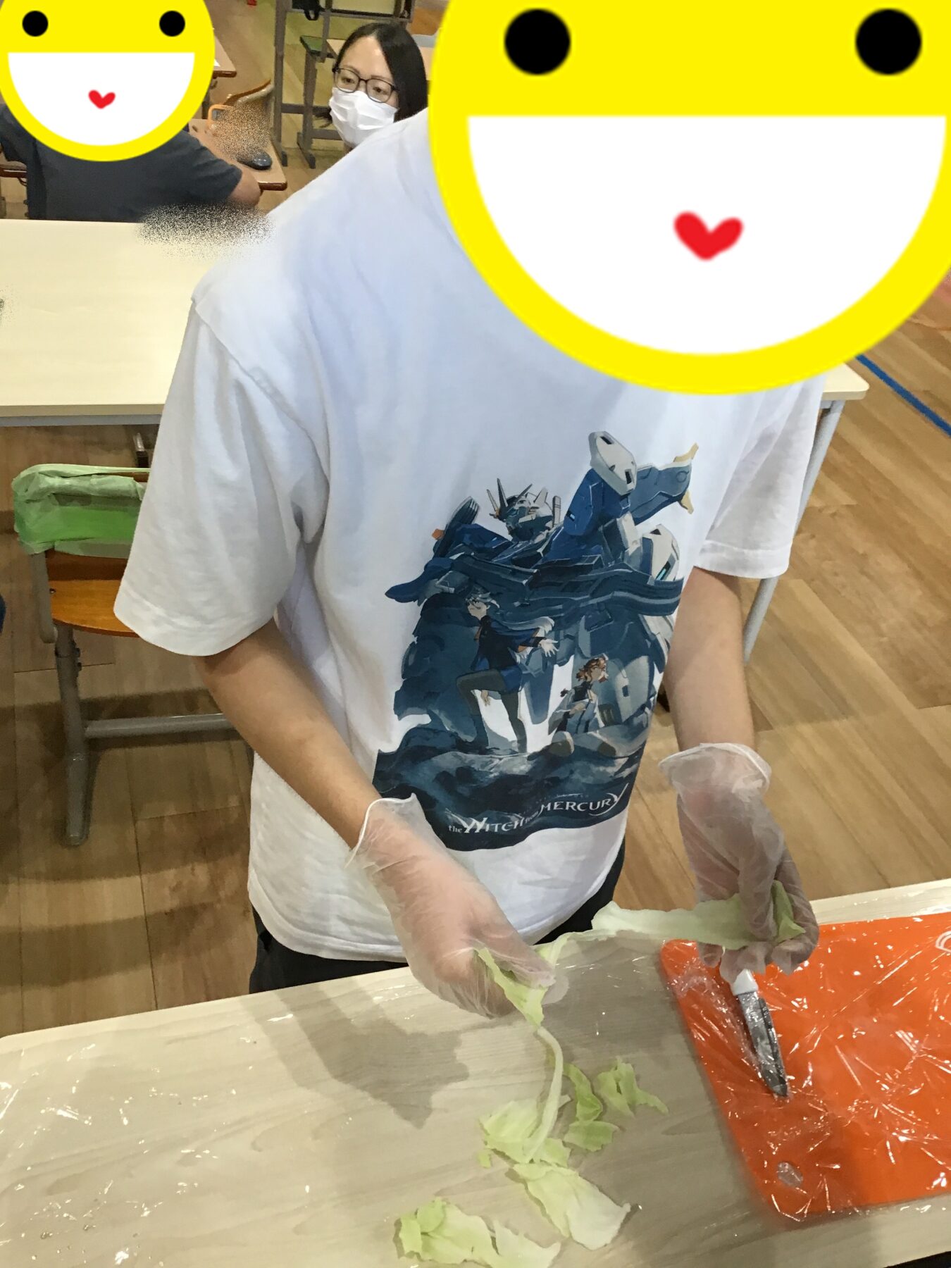 Person in a white graphic t-shirt wearing gloves chops lettuce at a table, with another masked person in the background.