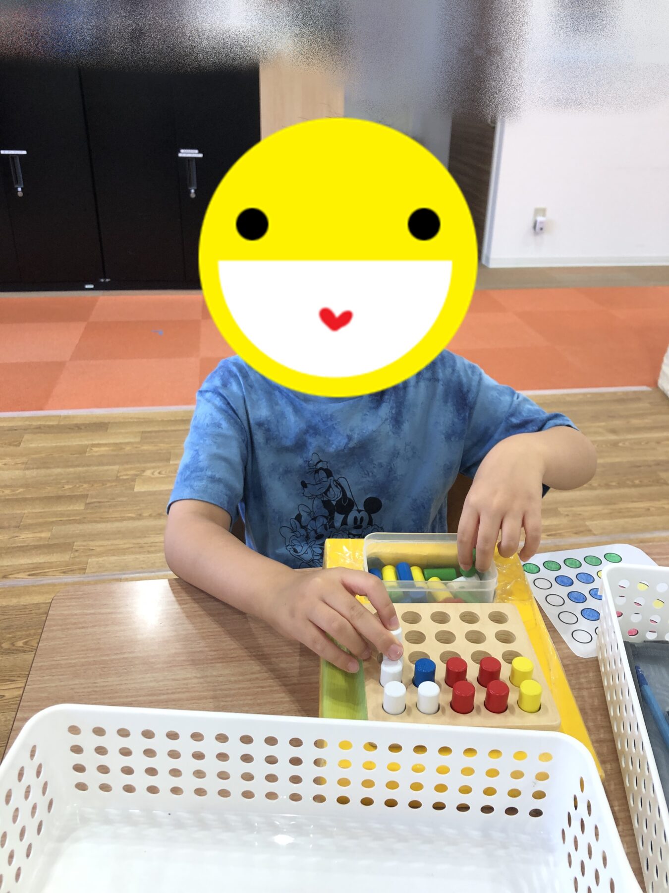 Child wearing a blue shirt sits at a table, playing with a wooden pegboard and colored pegs; face covered by a large yellow smiley sticker for privacy.