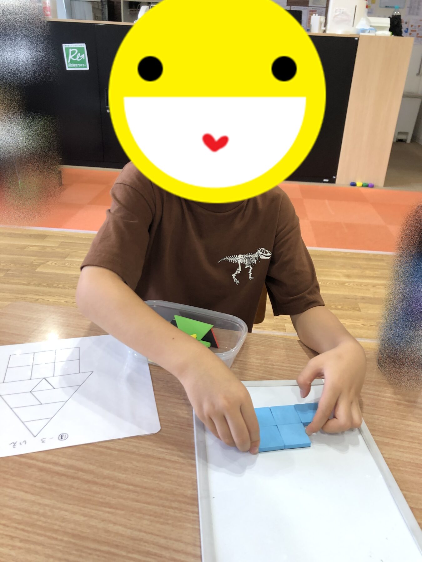 Child in a brown shirt builds a puzzle with blue tiles at a wooden table in a classroom, with papers and a plastic box nearby.