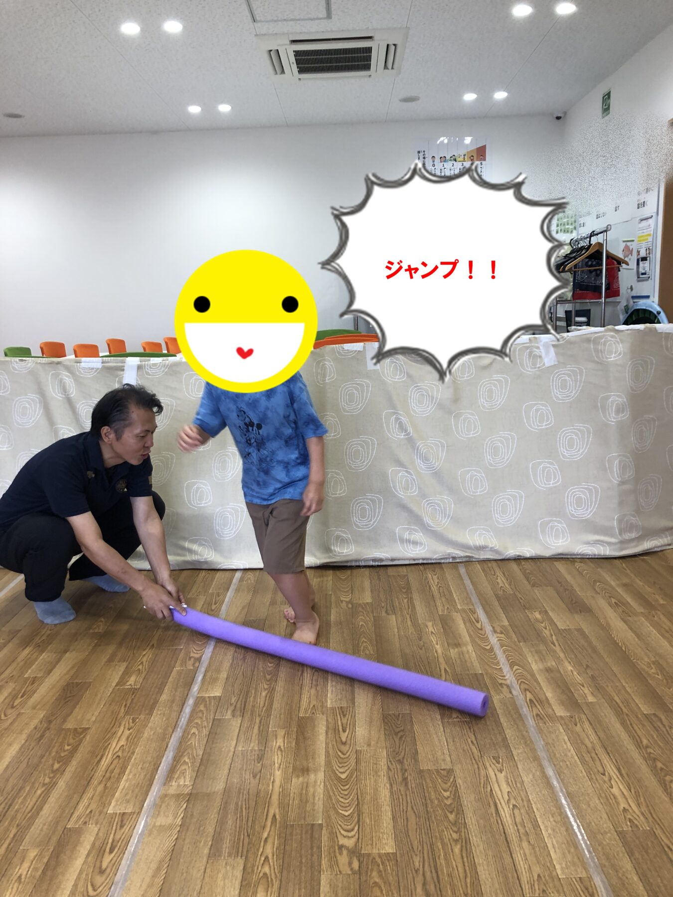 Child with a yellow smiley face covering their head, jumping with a purple pool noodle under supervision in a colorful indoor play area. Text in scene reads 'ジャンプ！！' (Jump!!)