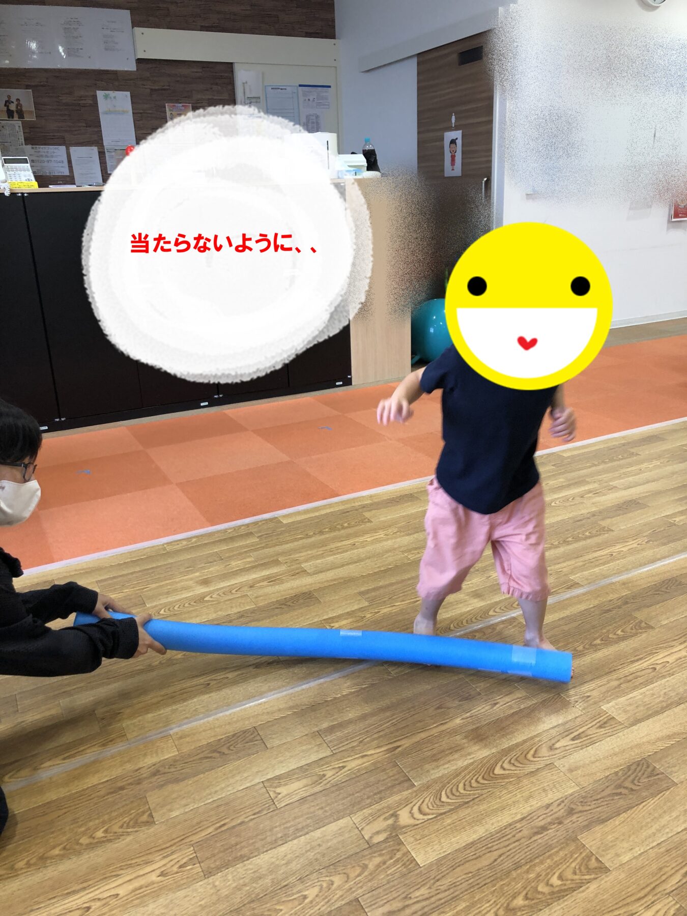 Young child in a navy shirt and pink shorts runs across a gym-like room toward a blue foam roller held by an adult on the left; the child’s face is covered by a large yellow smiley sticker.